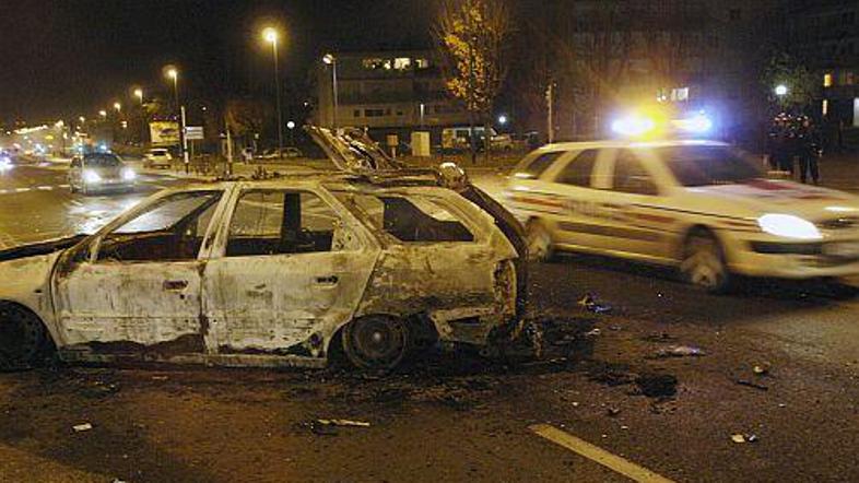 Policijski avtomobil se pelje mimo zažganih ostankov vozila, žrtve nočnega nasil