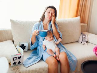 utrujena ženska sedi na kavču, pije kavo z dojenčkom v naročju.