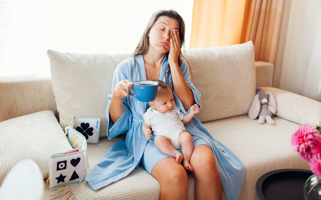 utrujena ženska sedi na kavču, pije kavo z dojenčkom v naročju.