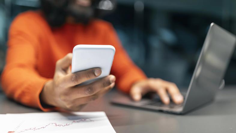 Moški iz Indije drži v roki telefon ob računalniku